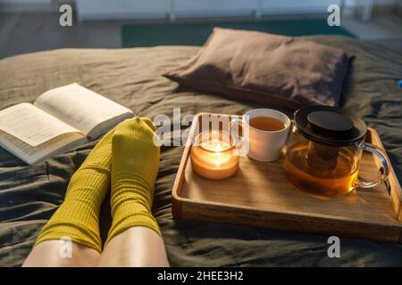 Soft photo of woman`s legs in the woolen socks on the bed with book and cup of tea and candle on the tray. Interior and home cosiness concept. Top vie Stock Photo